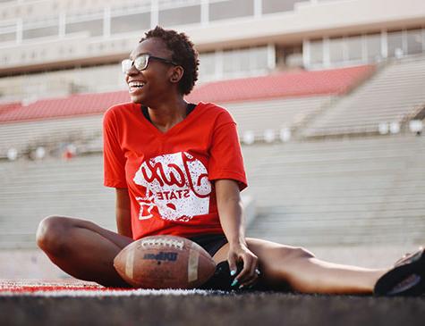 A female JSU student wearing a Jax State tee sits on the field at JSU stadium with a football in front of her